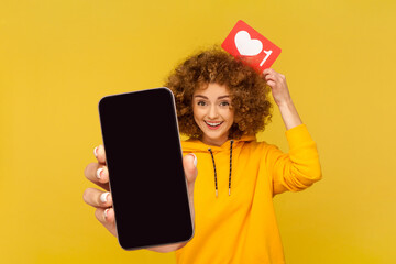 Funny positive woman blogger with Afro hairstyle wearing casual style hoodie showing mobile phone...