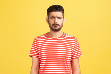 Fototapeta premium Portrait of serious sad upset man with beard wearing red T-shirt standing looking at camera, expressing sorrow and sadness, hearing bad news. Indoor studio shot isolated on yellow background.