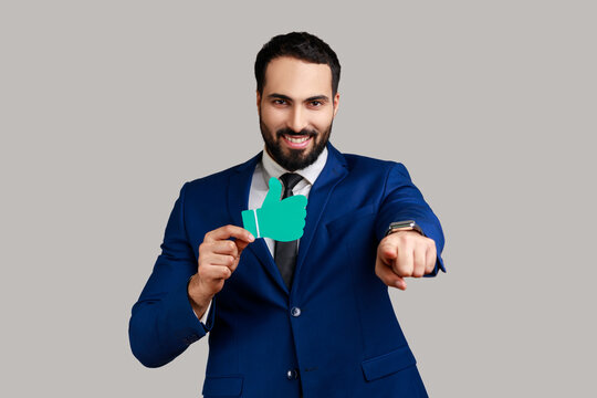Portrait of delighted handsome bearded businessman pointing at camera with smile and showing paper thumb up, wearing official style suit. Indoor studio shot isolated on gray background.