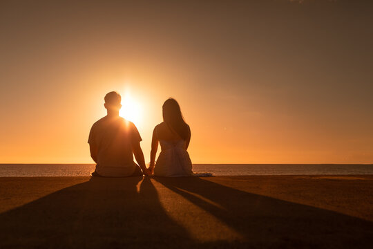 Couple holding hands at sunset facing the ocean sunset. . Love and relationships concept.	