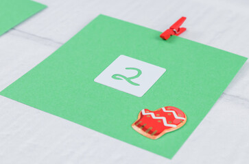 One green sheet of paper with numbers 2 and Christmas decor attached with a red mini clothespin to a white brick wall.