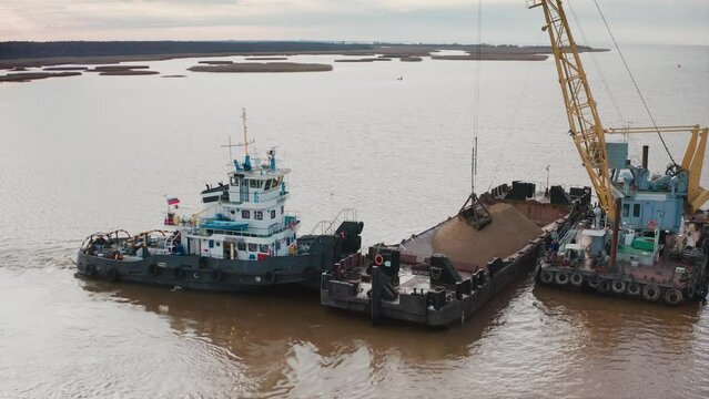 A floating barge with a pile of sand held by a tugboat. The bucket of the crane scoops the sand out of the pile. Aerial drone shot