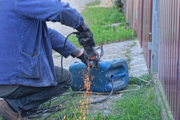 one worker in blue clothes cuts an iron rod with an electric grinder in the street in the green grass near the fence