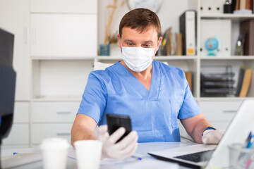 Fototapeta na wymiar Portrait of male doctor in face mask and gloves working on laptop consulting patient online, telemedicine concept