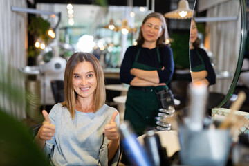 Portrait of cheerful young female client showing thumbs up satisfied with service in professional hairdressing salon.