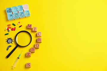 Word Microbes made with wooden cubes, pill box, syringe and magnifying glass on yellow background, flat lay. Space for text