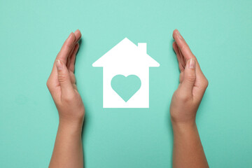 Woman holding hands around paper silhouette of house on turquoise background, top view. Insurance concept