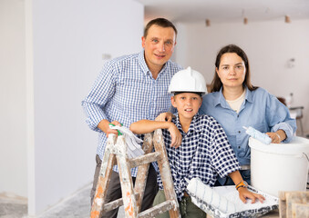 Portrait of positive family standing at stepladder in new apartment, holding paint rollers.