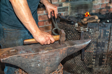 A forger wearing blue jeans and a t-shirt holds fire thongs and a metal twisted bolt on a vintage anvil. There's rounding hammer tool with a wooden handle in the blacksmith foundry shop with a furnace