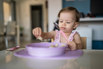 One child small caucasian toddler female baby eating at the table alone at home in room real people copy space early child development concept learning front view hold food healthy eating