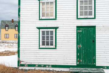 The exterior of a vintage wooden white house with three glass windows. The wooden cement windows have nine panes of glass and green decorative trim. The single green wooden door has a vintage latch. 
