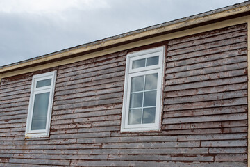 Two white vinyl windows with white trim and clear glass. The exterior wall has pine clapboard siding with red paint peeling. The boards are weathered and worn on the exterior of an old flat roof barn.
