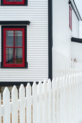 Exterior white wooden wall of horizontal clapboard siding on a vintage house with green trim. There's a small four pane glass window with red and green trim and a white picket fence in the foreground.