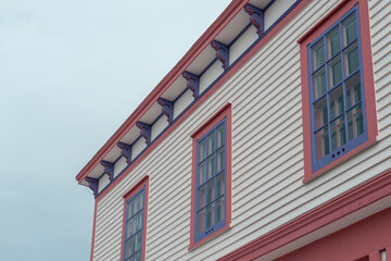The roof section of a large white vintage wooden building with decorative pink and purple wood trim. There are three multi-pane windows on the top floor. The background is a dramatic blue cloudy sky.