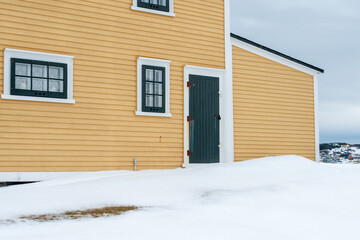 A bright and cheerful yellow colored country style house with wood cape cod clapboard, and white and black trim. There are multiple windows and a single black door. The ground is covered in snow.