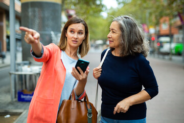 Friendly girl helping elderly woman to find necessary address in city, pointing direction with hand. - obrazy, fototapety, plakaty