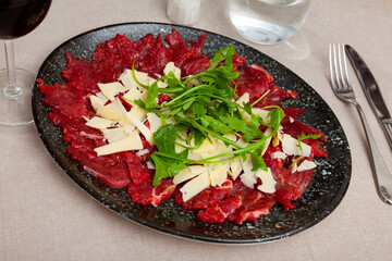 Raw beef meat, carpaccio, served on plate and topped with pieces of cheese and arugula.