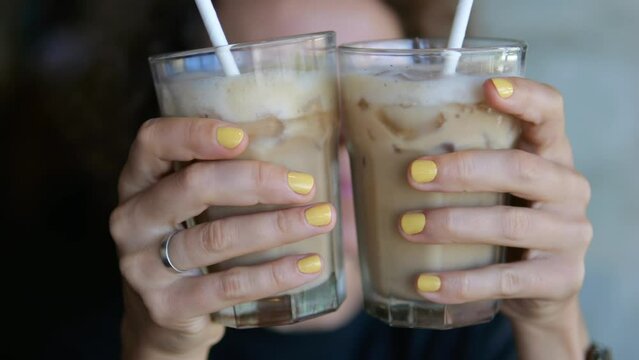In the foreground, close-up, a woman holds two glasses with a chocolate cocktail with milk and ice in her hands. She peeps out laughing, now from one side, then from the other.