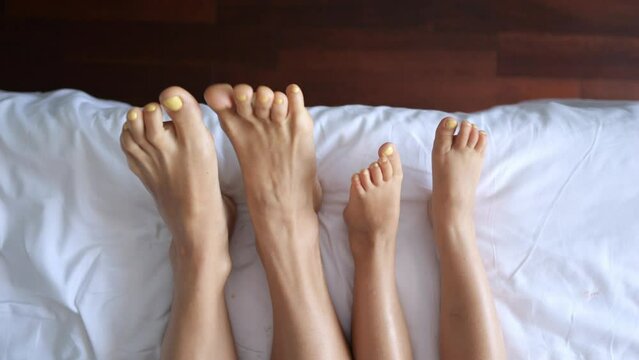 Close-up on top of two pairs of legs. A woman with a pedicure on her feet and a small child woke up in a hotel room on a snow-white bed. They wiggle their toes. Exercise helps wake up the brain.