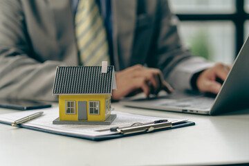Real estate office, sales representatives sitting at a desk with contract documents and a laptop and a small house next to it.