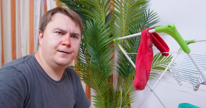 Young man with a red nose takes a clean sock off clothes drying rack, checks it, but finds a hole in it, front view. Guy sighs in frustration, shakes his head and leaves.