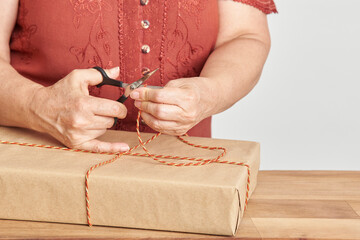 Obraz na płótnie Canvas Unrecognizable mature woman cutting a cord, tying a Christmas gift with a decorative string. Bright design with copy space. Concept of wrapping holiday presents.