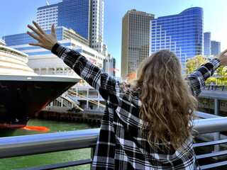 Fototapeta na wymiar a cheerful teen girl stands against the backdrop of Cruise ship Canada City Vancouver in a beautiful dress, she raised her hand, you can advertise a travel agency trip to Vancouver BC Canada