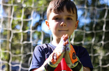 Menino jogando futebol na posição de goleiro, em frente às redes , mãos juntas com luvas 