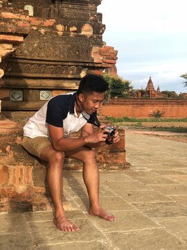 Young man holding a camera and sitting near ancient site in Nyaungu, Mandalay Region, Myanmar (Burma)