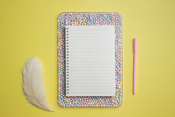 White notebook over the colorful ball with pink pen and white feather on yellow background. 