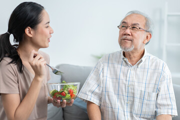 Young Asian daughter serving and feeding food to senior father while he using and playing on digital tablet in the living room. family love and support to elderly people. 