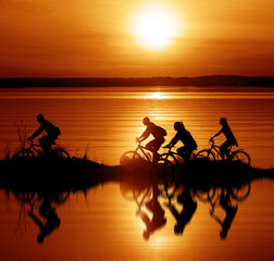 Fototapeta na wymiar Image of sporty company. friends on bicycles outdoors against sunset sky. Silhouette of motion go of four 4 cyclist along shoreline coast. Reflection sun on water. Copy Space for inscription