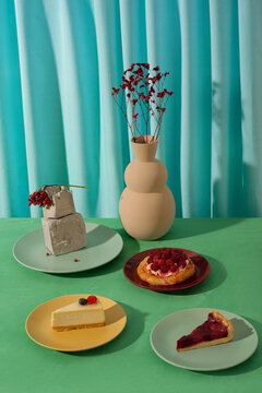 Vertical high angle still life studio composition of pies, cakes, gypsum decor element and vase with flowers on green table against blue background