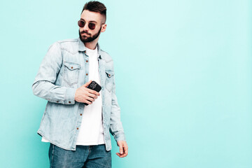Fototapeta na wymiar Portrait of handsome confident stylish hipster lambersexual model.Man dressed in jacket and jeans. Fashion male posing near blue wall in studio. Holding smartphone