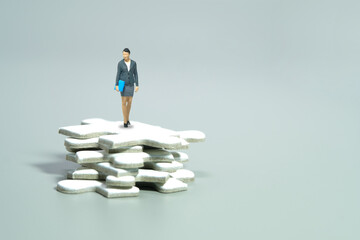 Miniature people toy figure photography. A businesswoman standing above puzzle jigsaw pile. Isolated on grey background