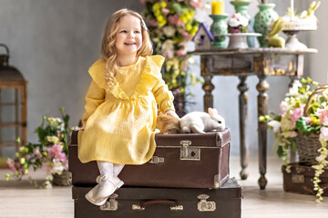A little blonde toddler girl in a yellow dress sits on old suitcases with live rabbits. Emotions....