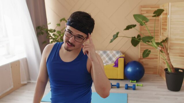 Funny man with glasses, moustache and goatee in bright blue retro sportswear tank top tapping his head thinking. Be smart. Positive motivation exercise indoors sport equipment in the background