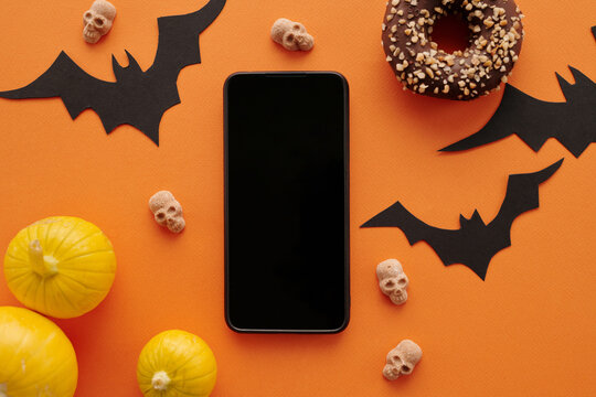 Top-down view shot of flat lay composition with smartphone, paper bats, pumpkins, donut and small toy skulls on orange background
