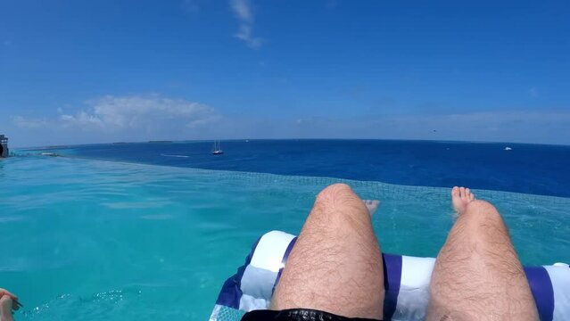 Man relaxing in the pool, stock video. A guy floating in a hammock in an infinity pool overlooking the ocean at a luxury hotel in the Maldives. Wide angle shot.