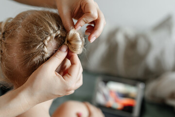 Mom's hands weave a braid on the head of a blonde daughter. Close-up. Mother and child spend time...