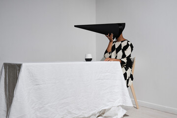 Horizontal studio shot of mysterious young African American woman wearing black and white dress with creepy conus-shaped mask on face sitting at table