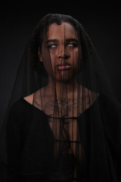 Vertical waist up studio portrait of creepy African American woman wearing black dress with veil and blind white lenses for Halloween party standing in darkness