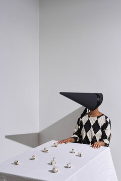 Vertical conceptual shot of unrecognizable woman wearing black and white outfit with conus-shaped mask on face sitting at table with cotton flowers on it