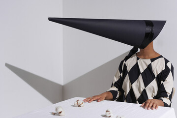 Horizontal medium shot of modern young African American woman wearing black and white outfit with creepy conus-shaped mask on face sitting at table with cotton flowers on it