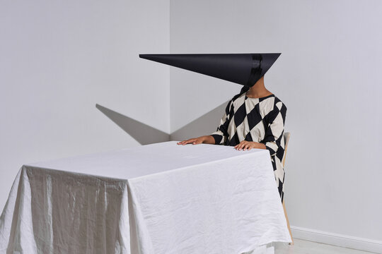 Horizontal studio shot of modern young African American fashion model wearing black and white outfit with creepy conus-shaped mask on face sitting at table