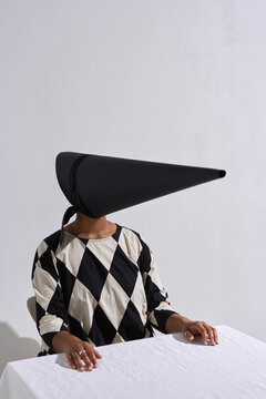 Vertical studio portrait shot of unrecognizable young African American fashion model wearing black and white outfit with creepy conus-shaped mask on face sitting at table