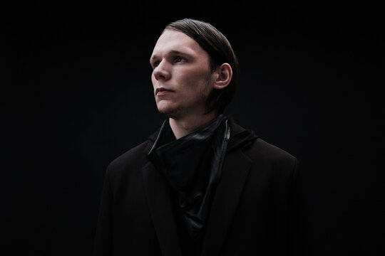 Horizontal medium studio portrait of stylish young Caucasian man wearing dark fashionable outfit standing against black background looking away