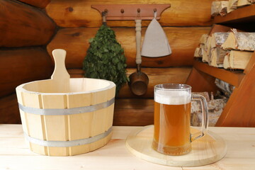 A mug of light beer stands on a boards desk near the wooden vat for the sauna on the background a...
