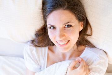 Obraz na płótnie Canvas Dreamy girl lying on white bed with large pillows, hugging blanket with her arms and smiling with white teeth looks at camera, not wanting to wake up quickly, enjoying her free time on the weekend