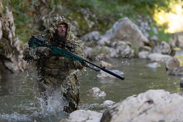 A military man or airsoft player in a camouflage suit sneaking the river and aims from a sniper rifle to the side or to target.